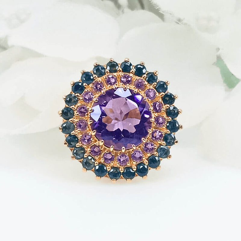 Close-up view of the Floral Radiance Ring, featuring a round silver ring plated with gold and a floral design made up of sapphire and amethyst gems surrounding a central violet amethyst.