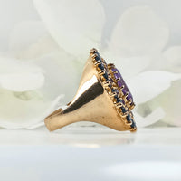 featuring a round silver ring plated with gold and a floral design made up of sapphire and amethyst gems surrounding a central violet amethyst.